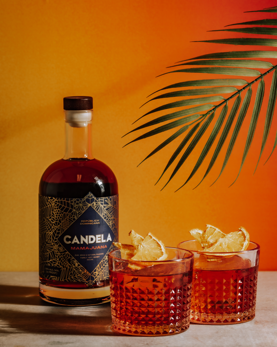Negroni Recipe with spiced rum, called Mamajuana. Image shows bottle of Candela Mamajuana, a Dominican Spiced Rum with the cocktails. The Negroni cocktail is served over large ice cubes, with dried fruit, and in a rocks glass.
