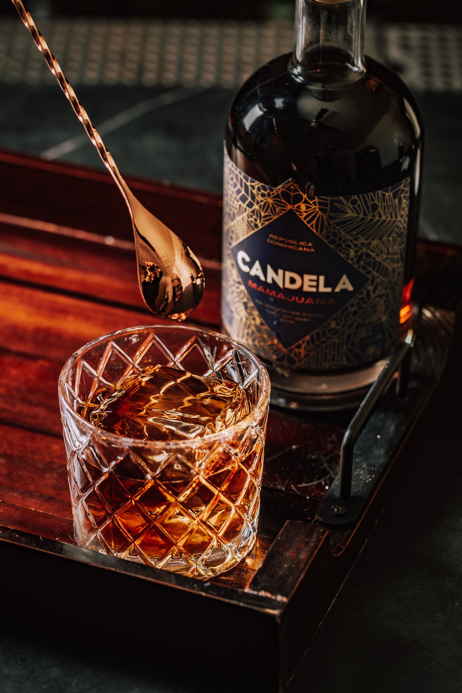 The Sugar Daddy Cocktail by Candela Mamajuana. This cocktail recipe is a Dominican take on the classic Old-Fashioned, and uses spiced rum. Image shows cocktail over a large ice cube, stirred, in a rocks glass next to a bottle of Candela Mamajuana.
