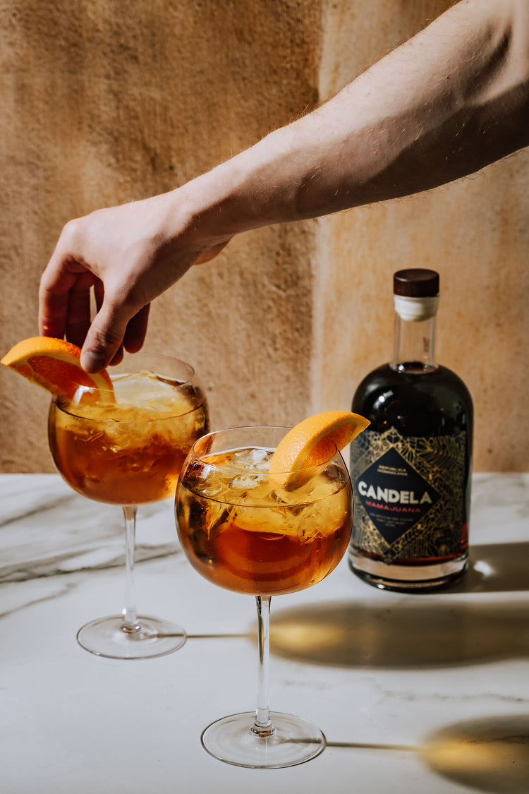 The Dominican Spritz – a spiced rum cocktail prepared with Candela Mamajuana. The cocktail recipe is a Dominican take on the traditional Aperol Spritz. Image features 2 Dominican Spritz Cocktails with fresh orange and a bottle of Candela Mamajuana